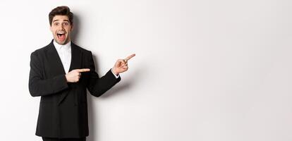 Image of attractive smiling guy dressed for new year party, pointing fingers right and showing advertisement, looking impressed standing over white background photo