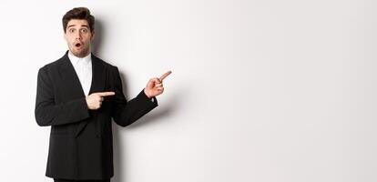 Portrait of surprised handsome man with beard, wearing black suit, open mouth and pointing fingers right at advertisement, standing over white background photo