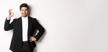 Concept of new year party, celebration and lifestyle. Portrait of confident, successful businessman in suit, showing okay sign and winking, approve something good, white background photo