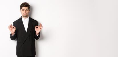 Concept of new year party, celebration and lifestyle. Portrait of confident good-looking man in black suit, showing okay sign and approve something, standing over white background photo