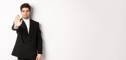 Portrait of serious handsome man in formal suit, extending hand to stop you, prohibit action, forbid and disagree with something, standing against white background photo