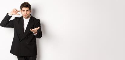Portrait of confused handsome businessman scolding employee, pointing at head and camera, standing disappointed over white background photo