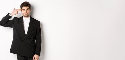 Portrait of distressed and troubled businessman in suit, pointing finger gun at head and looking bothered, shooting himself, standing against white background photo