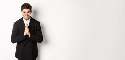 Portrait of handsome man in black suit, being grateful, saying thank you and bowing politely, smiling as holding hands together, expressing gratitude, standing over white background photo