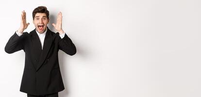 Frustrated and concerned man in black suit, screaming in panic and looking at something shocking, standing over white background photo