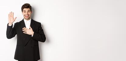 Image of handsome smiling guy telling the truth, raising on arm to promise something, standing over white background in black suit photo