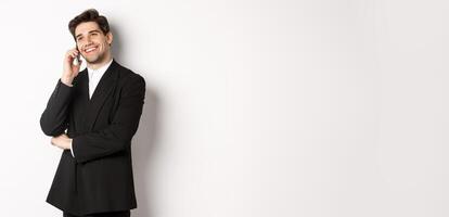 Image of handsome and successful businessman talking on phone, smiling pleased, standing in suit against white background photo