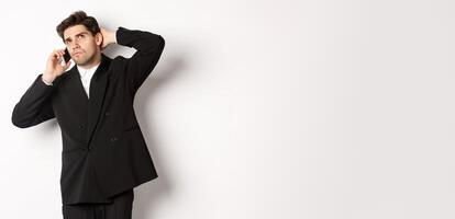 Image of indecisive businessman talking on phone and thinking, looking doubtful, making decision, standing over white background photo