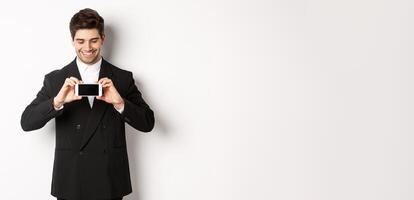 Portrait of handsome bearded man in black suit, showing you smartphone screen and smiling pleased, standing against white background photo