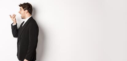 Profile shot of handsome businessman in black suit talking on speakerphone, smiling and looking happy, recording voice message, standing over white background photo