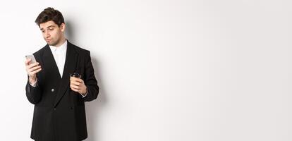 Portrait of thoughtful handsome businessman, drinking coffee and browsing in internet, looking at smartphone screen, standing against white background photo