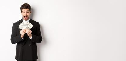 Image of handsome bearded guy in black suit, looking at money with excitement, standing over white background photo