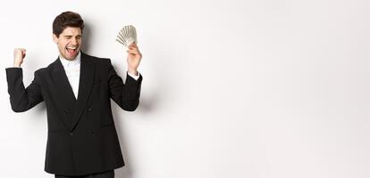 Portrait of handsome and successful businessman in suit, dancing with money, standing against white background photo