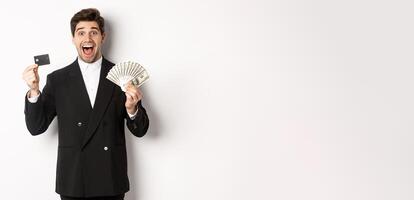 Image of handsome male entrepreneur in suit, showing credit card and money, smiling amazed, standing over white background photo