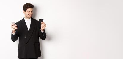 Portrait of confident handsome businessman shopping online, holding credit card and smartphone, standing over white background photo
