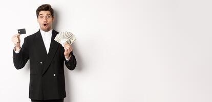 Portrait of surprised handsome man i suit, showing credit card with money, standing against white background photo