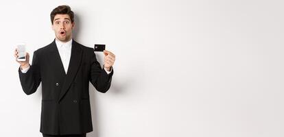 Image of handsome businessman in black suit, looking amazed and showing credit card with mobile phone screen, standing against white background photo