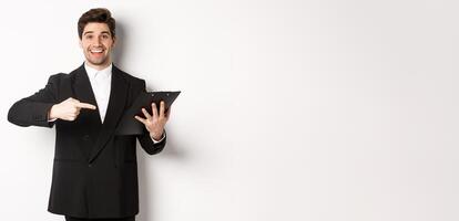 Image of handsome smiling businessman in black suit, pointing finger at clipboard with documents, standing against white background photo