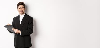 Image of handsome, successful boss in black suit, winking and smiling while signing documents, standing against white background photo