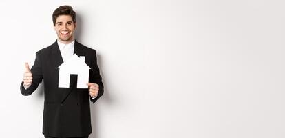 Portrait of confident real estate agent showing house maket and thumb-up, standing against white background photo