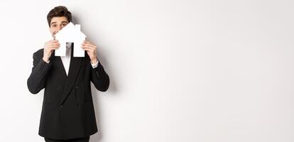Image of handsome broker in black suit, showing house maket and smiling, selling homes, standing against white background photo