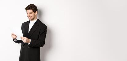 Image of attractive businessman in black suit, reading document and smiling, working on report, standing against white background photo