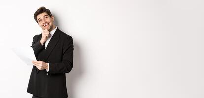 Image of thoughtful, handsome businessman in black suit, holding document and looking at upper right corner, thinking and smiling, standing against white background photo