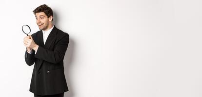 Image of handsome businessman in black suit, looking through magnifying glass, found something and smiling, standing over white background photo