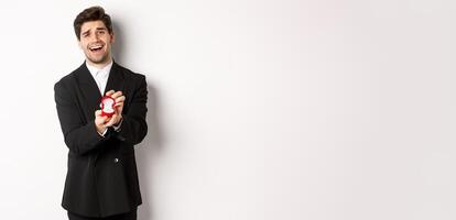 Portrait of handsome man in black suit, open box with wedding ring, making a proposal, asking to marry him, standing against white background photo