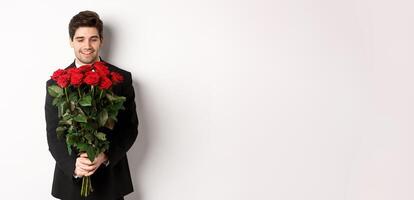Image of handsome boyfriend in black suit, holding bouquet of red roses and smiling, being on a date, standing over white background photo