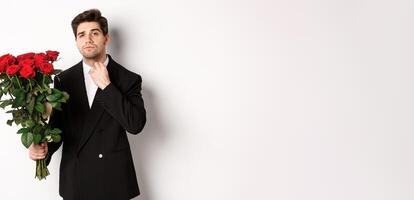 Image of elegant and sassy man in black suit, looking confident and holding bouquet of red roses, going on a romantic date, standing against white background photo