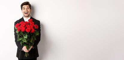 Image of handsome boyfriend in black suit, holding bouquet of red roses and smiling, being on a date, standing over white background photo