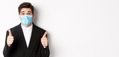 Concept of covid-19, business and social distancing. Close-up of happy businessman in black suit and medical mask, showing thumbs-up, making a compliment, white background photo