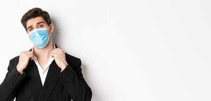 Concept of covid-19, business and social distancing. Image of confident handsome man in trendy suit and medical mask, looking sassy, standing against white background photo