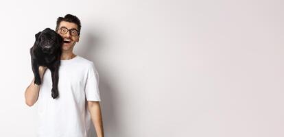 Cheerful hipster guy in glasses and t-shirt, carry cute black pug on shoulder and smiling. Dog owner playing with his pet, standing over white background photo