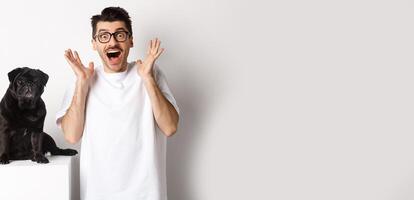 Ecstatic young man looking with excitement and rejoice, standing near cute black pug, staring at camera happy, standing over white background photo
