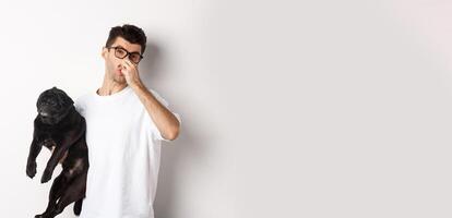 Young man shut nose as holding pug, disgusted with bad smell fart of animal, standing over white background photo