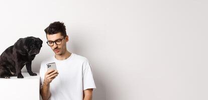 Young man showing photos on smartphone to cute dog, pet owner and pug staring at mobile phone, white background