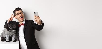 Cheerful dog owner in suit celebrating New Year with dog, taking selfie on smartphone near cute black pug in costume, white background photo
