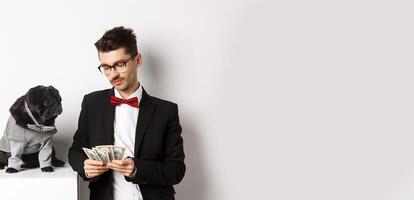 Handsome young man in suit standing near black pug in costume and counting money, working on parties, posing over white background photo