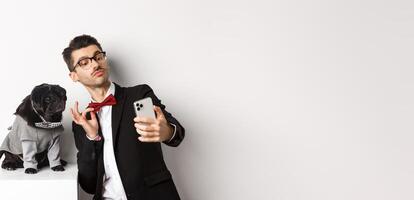 Cheerful dog owner in suit celebrating New Year with dog, taking selfie on smartphone near cute black pug in costume, white background photo
