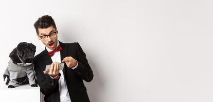 Amazed man showing to dog, pointing at mobile phone and look excited while pug sitting unbothered in costume, white background photo