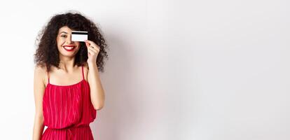 Image of elegant woman in red dress, showing plastic credit card and smiling at camera, standing over white background photo