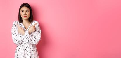 Concerned asian woman having doubts, pointing sideways and looking left with hesitant and sad face, need help with choice, standing over pink background photo