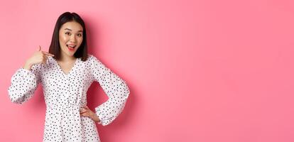 Beautiful asian girl in dress looking happy, pointing finger at herself, standing on romantic pink background photo