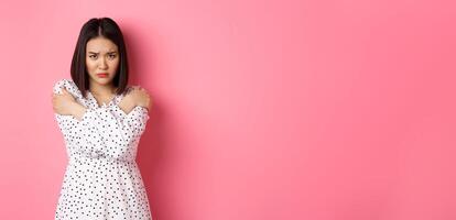 Timid and offended asian girl cross arms on chest, staring defensive and insulted at camera, standing in dress over pink background photo