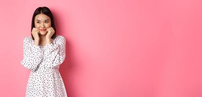 Cute and shy asian girl blushing, touching cheeks and looking left at copy space silly, standing against pink background photo