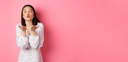 Beautiful chinese girl in dress blowing air kisses, sending mwah at camera, standing over pink background photo