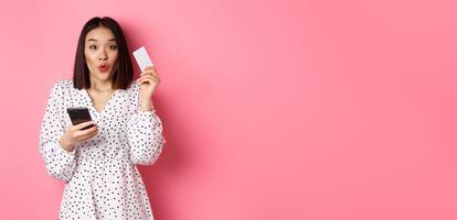 Attractive young asian woman order online, holding credit card and mobile phone, making internet purchase, standing happy over pink background photo