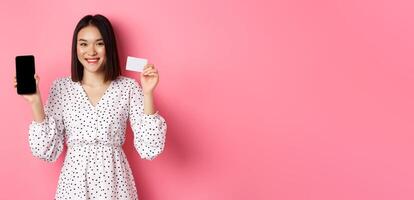 Cute asian woman shopping online, showing bank credit card and mobile screen, smiling and looking at camera, standing over pink background photo